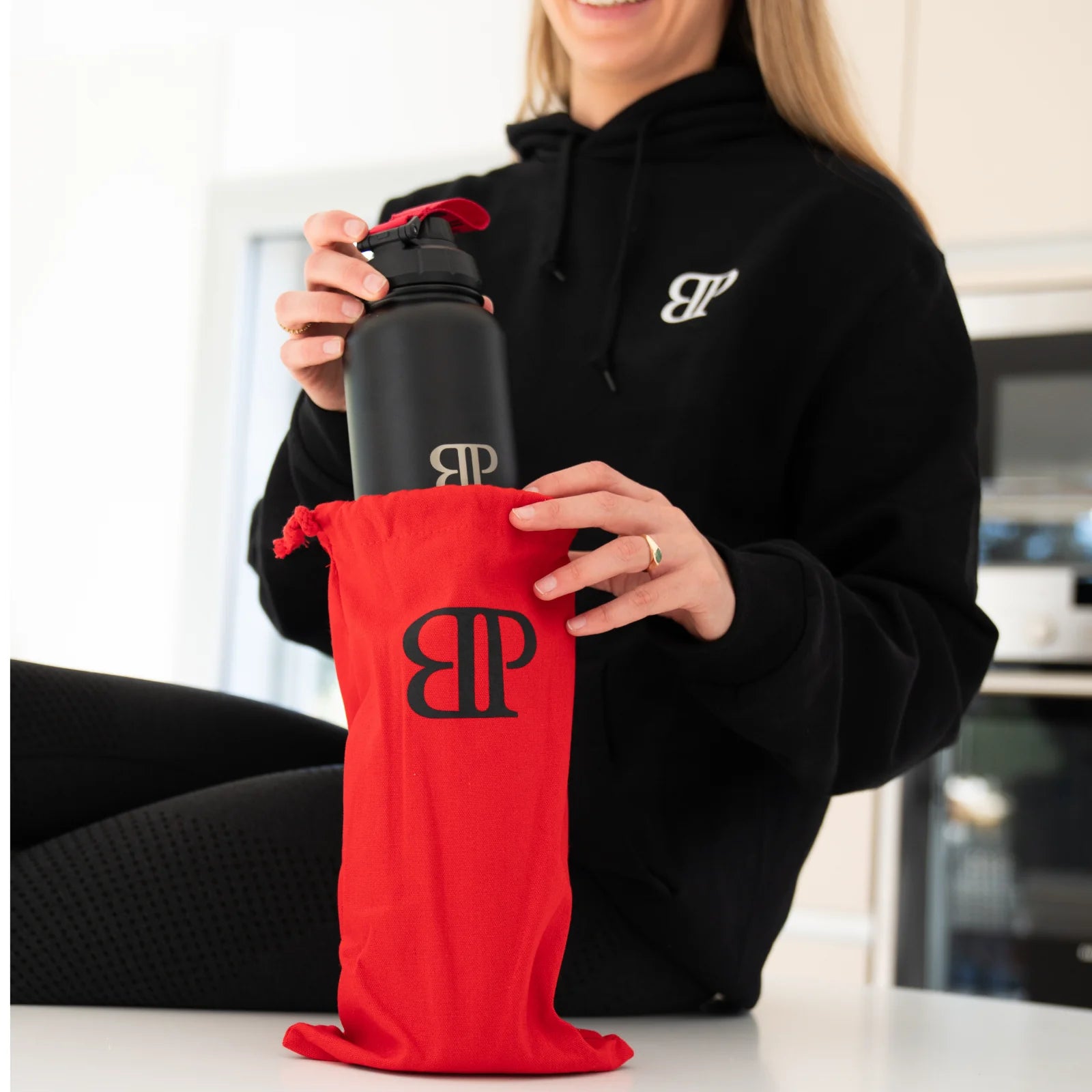 Frau zieht einen schwarzen Bollaz Thermo Shaker mit BP Logo aus einer roten Stofftasche, hochwertige Premium-Verpackung und stilvoller Hoodie.