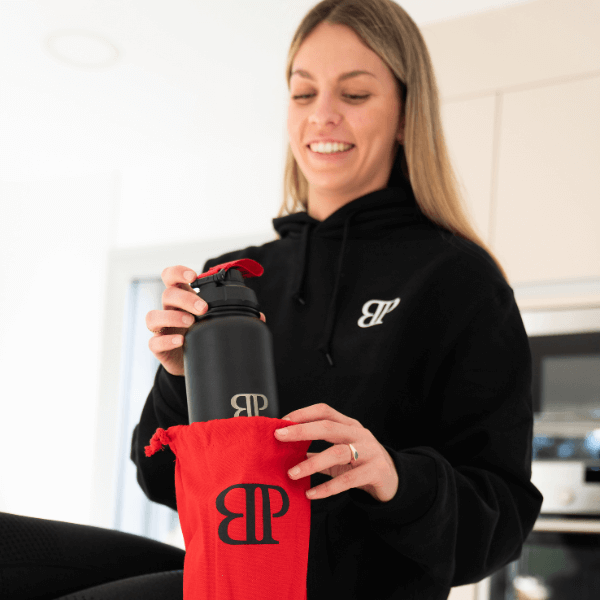 Frau mit schwarzem Bollaz-Hoodie packt schwarzen Bollaz Thermoshaker aus rotem Beutel mit BP-Logo in einer hellen Küche aus.